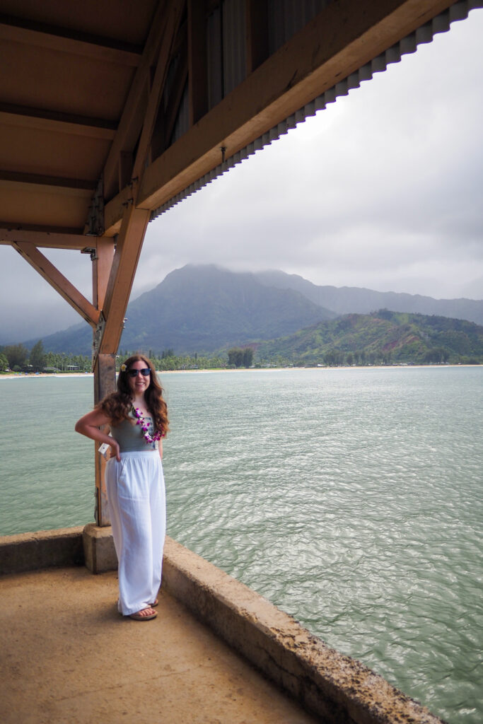 author, a young woman, standing on a pier in Hanalei with mountains and ocean behind her in the background