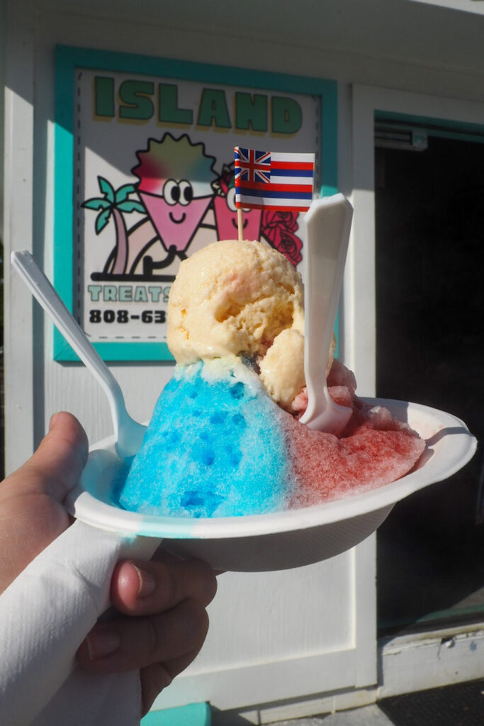 hand holding a bowl of shave ice, half blue, half red, with a Hawaiian flag stuck in it