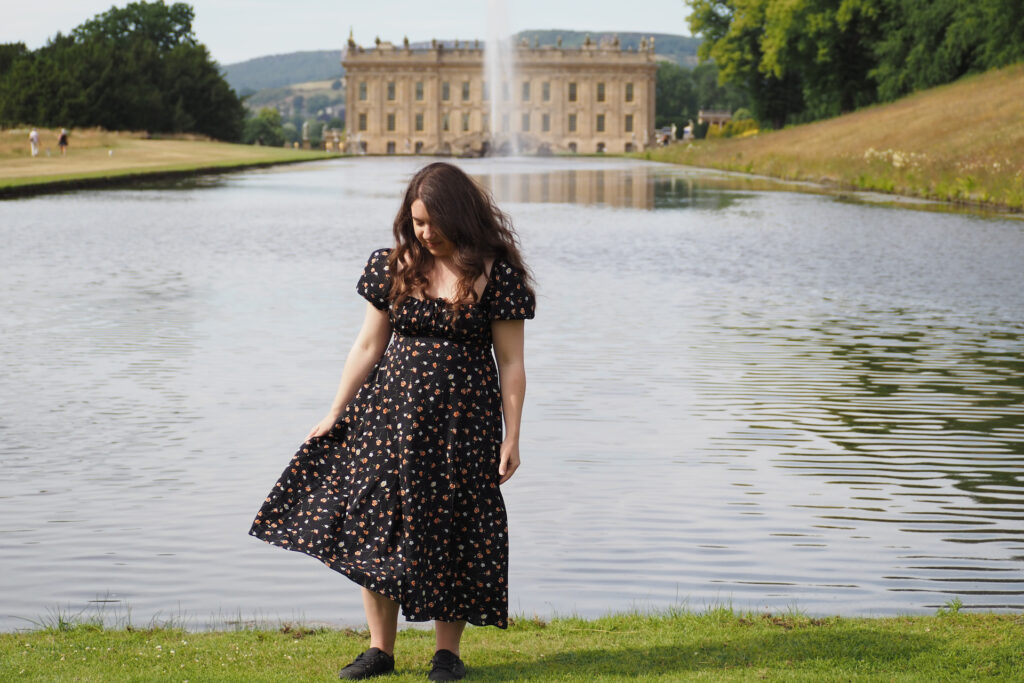 girl in a black dress standing in front of a pond with a fountain and a mansion in the background