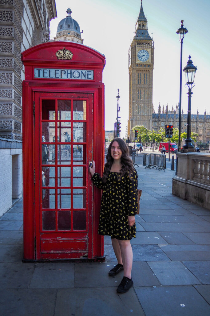 A woman in a black dress with yellow patterns stands next to a classic red telephone booth in London. Behind her are Big Ben and the Palace of Westminster on a sunny day.