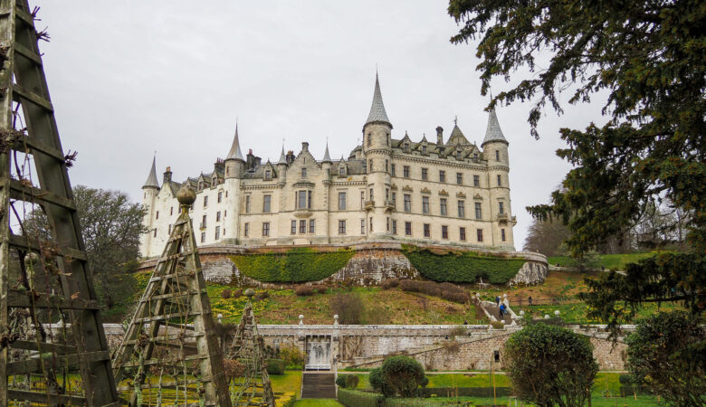 A grand white stone castle with multiple turrets sits on a hill, surrounded by lush green gardens, hedges, and trellises under a cloudy sky. Paths and stone walls lead up to the historic building.