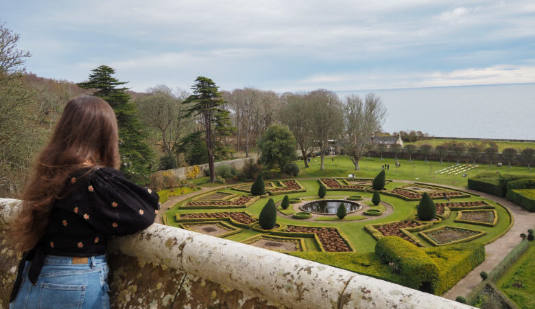 A woman with long brown hair, wearing a black top and jeans, stands at a stone balcony overlooking an ornate formal garden with geometric hedges, topiary, and a central fountain near the coastline.