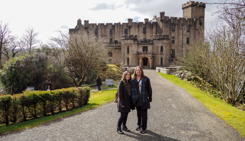 Two women in dark coats stand on a gravel path leading to a large stone castle with towers, battlements, and a flag on top. Trees and bushes line the path, and a few people are in the background near the castle.