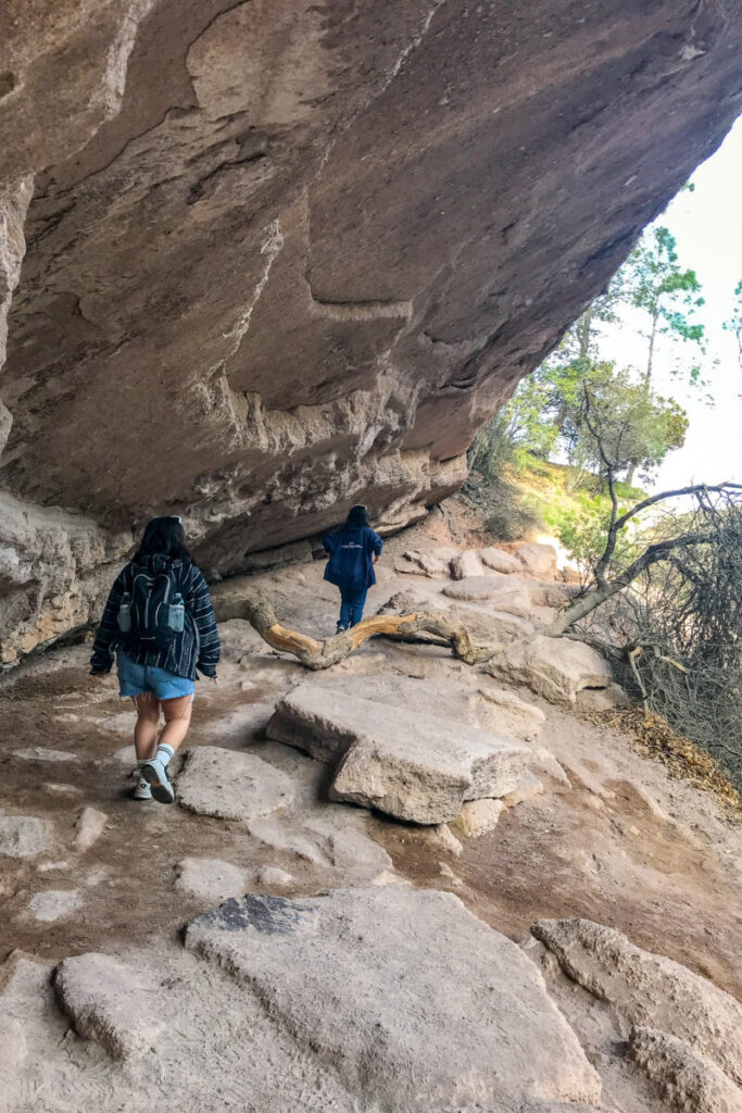 Two people with backpacks hike along a rocky trail beneath an overhanging cliff, surrounded by sparse trees and sunlight filtering through branches.