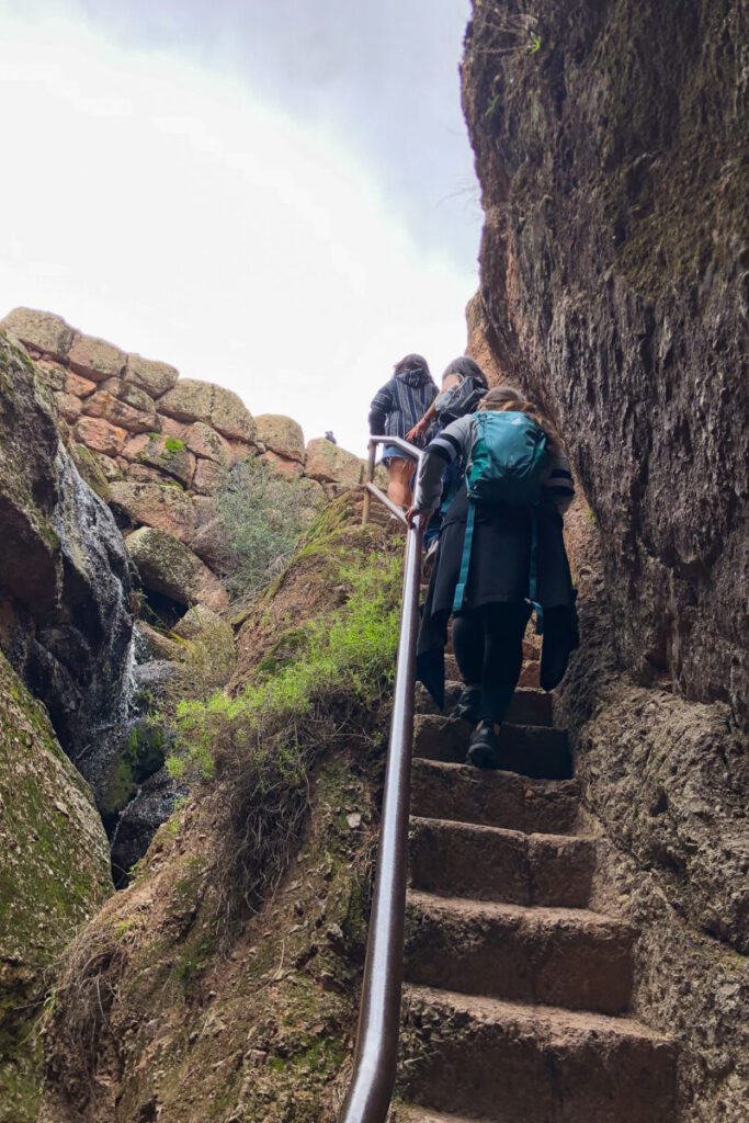 Three people climb a narrow stone stairway between large rock walls, using a metal handrail. One wears a teal backpack. Green moss and a small waterfall are visible on the left side.