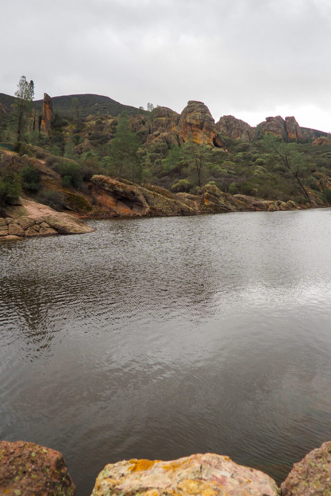 A calm lake with rocky hills and sparse green trees under a cloudy sky. The rocks have orange and brown hues, and the landscape appears rugged and natural.