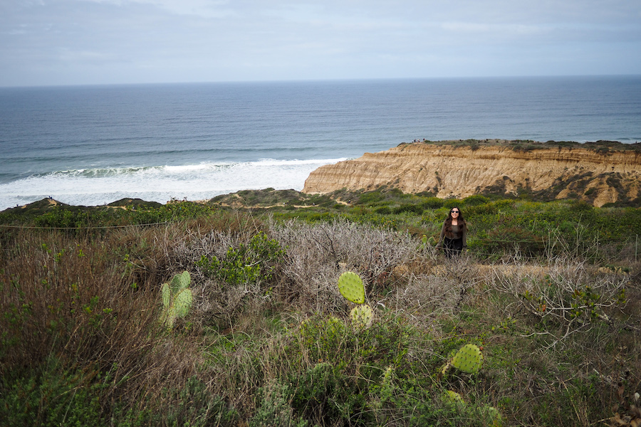 girl wearing a green shirt standing on a trail by a cliff and the ocean