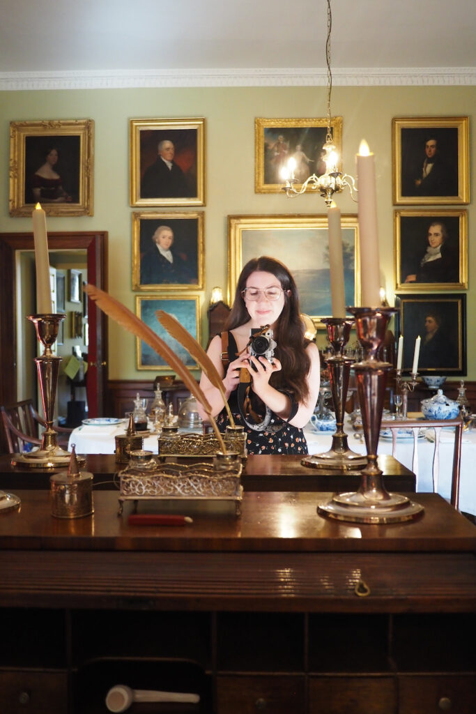 A woman takes a mirror selfie in an elegant room with vintage decor, tall candles, and framed portraits on the wall behind her.