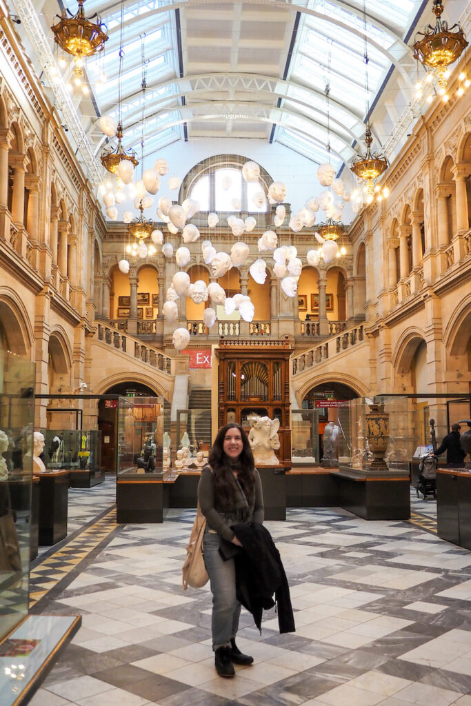 A woman stands smiling in the center of a grand hall with arched balconies, marble floors, and glass display cases, under a ceiling with hanging white head sculptures and ornate chandeliers.
