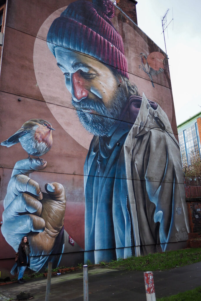 author, a young woman, standing by the St Mungo mural, which is a man looking at a bird sitting on his finger