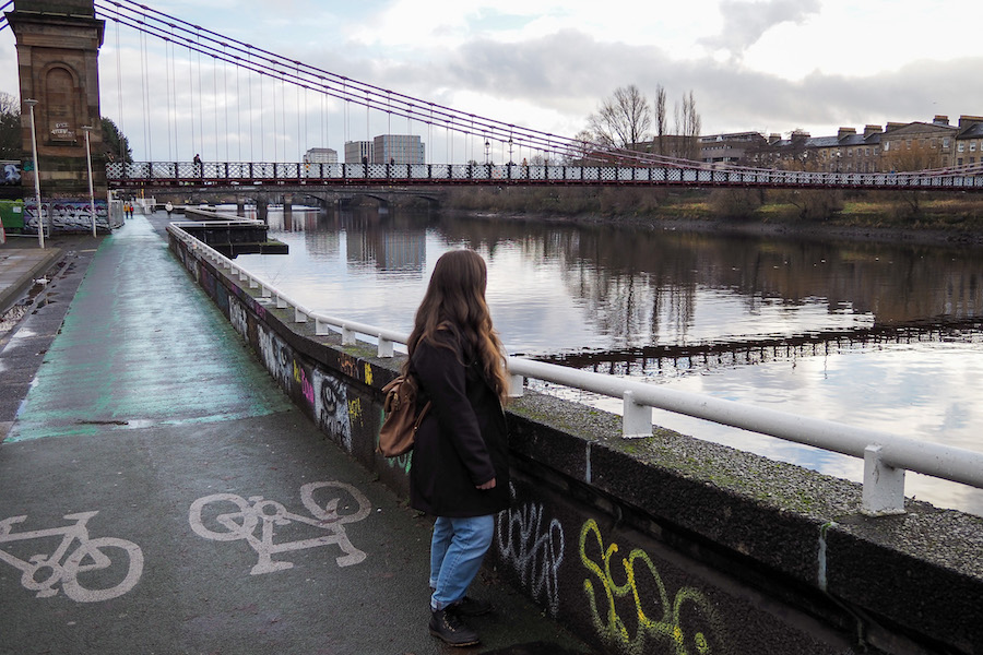 A person with long hair and a backpack stands by a river, looking at a suspension bridge. Bicycle path markings and graffiti are visible on the riverside path. Buildings and trees line the opposite bank.