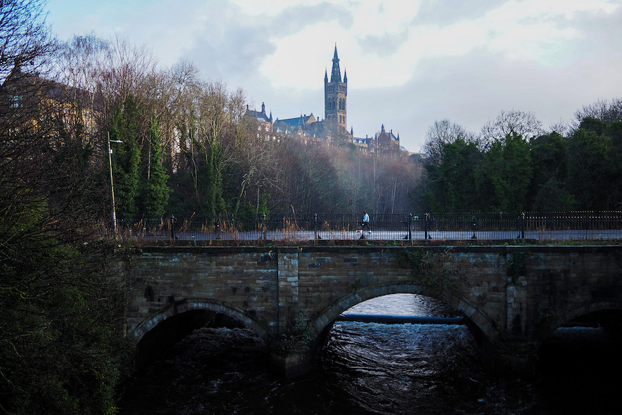 A stone bridge spans a river beneath dense trees, with a person walking along the path above. In the background, a tall, gothic-style tower rises above the treetops under a cloudy sky.