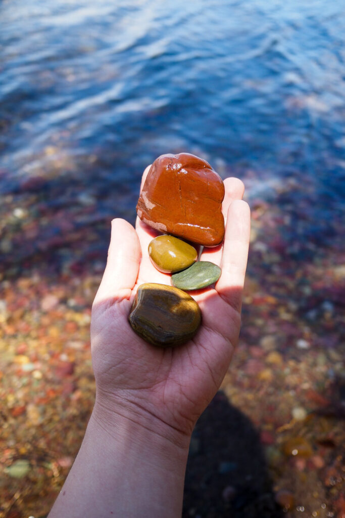 a hand holding colorful pebbles in front of a lake with clear water in Glacier National Park