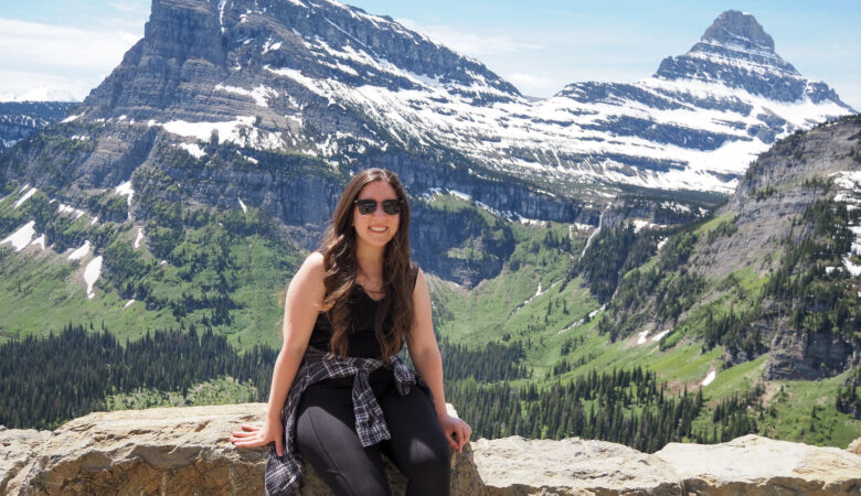 girl sitting on a wall at an overlook at Glacier National Park with snow-topped mountains behind her. Part of 2 day itinerary in Glacier National Park