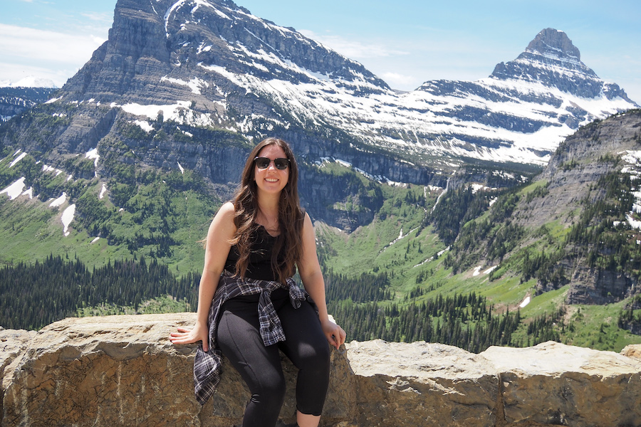 a woman wearing sunglasses sitting on a wall at an overlook on the Going-to-the-Sun Road in Glacier National Park