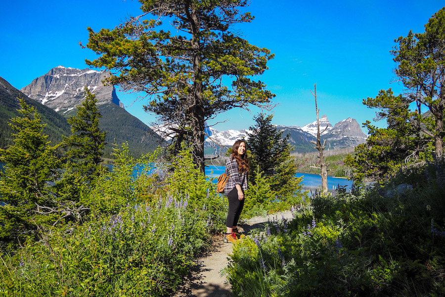 a woman standing on a trail with purple wildflowers lining the trail and a lake in the background