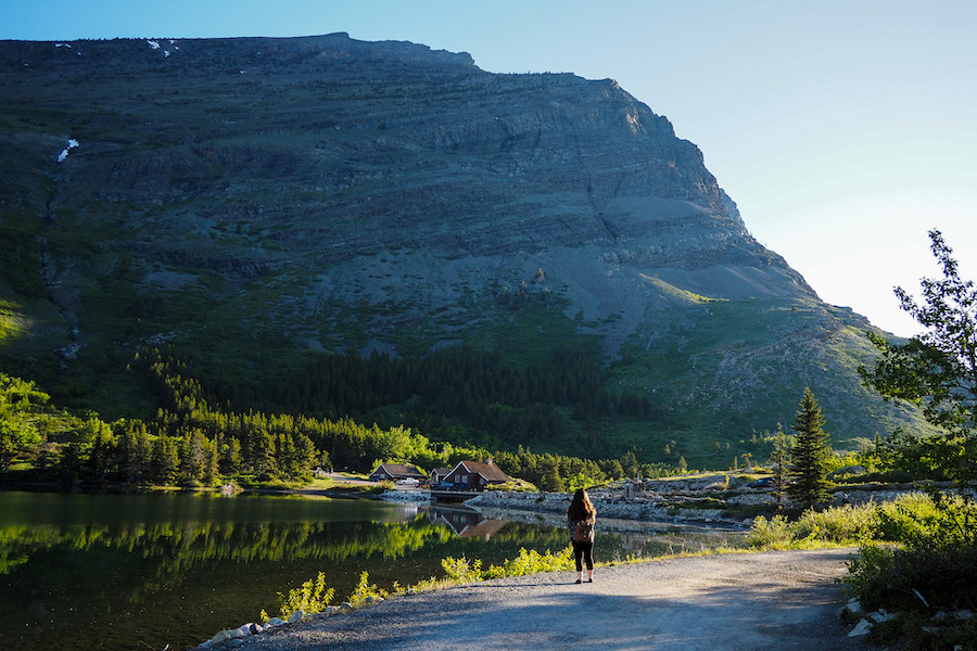 a woman standing on a rocky road looking out at the view of the lake in Many Glacier