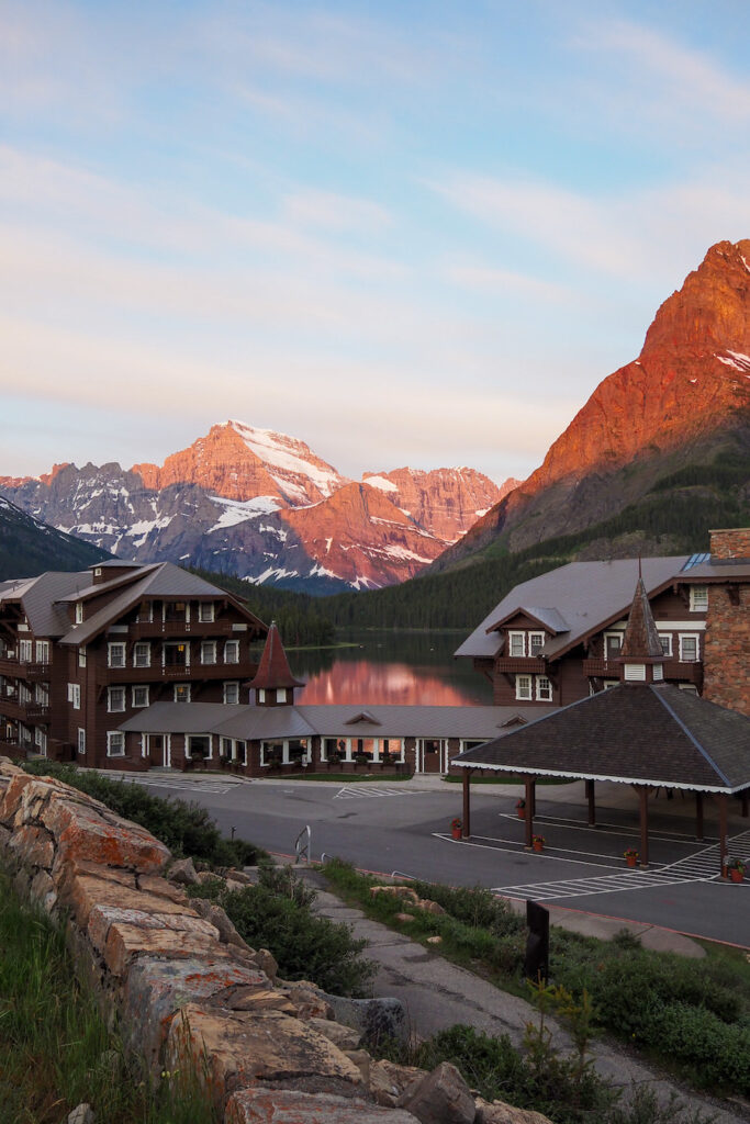 a sunrise making mountains colorful with a reflection on the lake in Glacier National Park with the Many Glacier Hotel in front