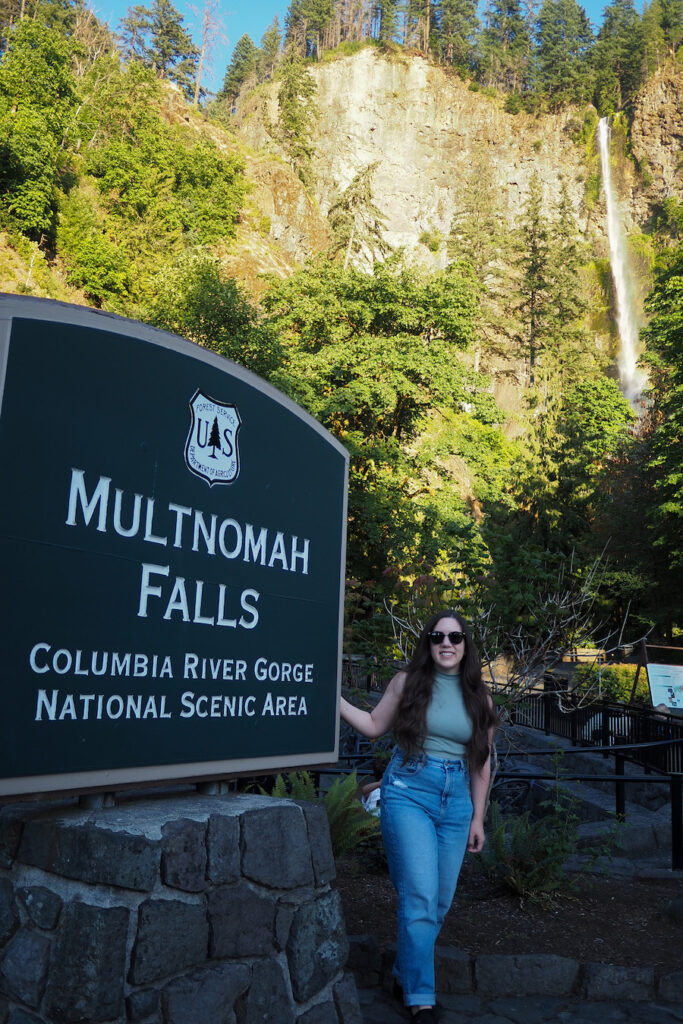 a girl standing next to a sign that says "Multnomah Falls: Columbia River Gorge National Scenic Area" with a waterfall in the background