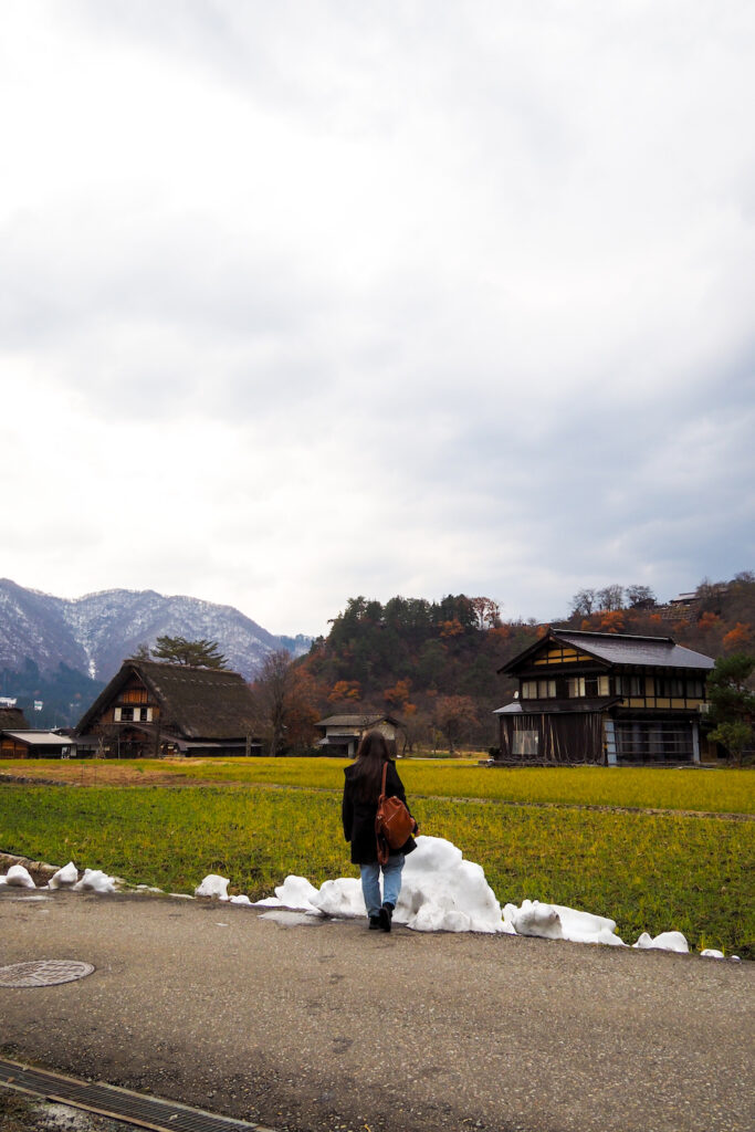 girl standing on a road near a pile of snow with traditional houses in the background in Japan