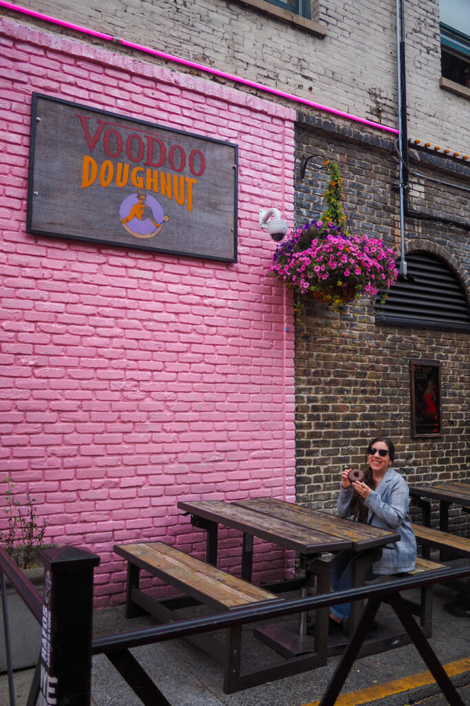 a girl sitting on a brown bench holding a chocolate donut with a pink brick wall behind them, and a sign on it that says "Voodoo Doughnut" in Portland