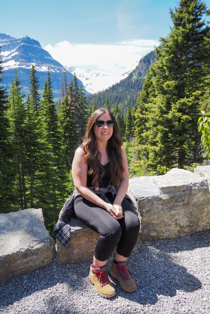 a woman wearing sunglasses sitting on a wall in front of the view of Jackson Glacier in Glacier National Park