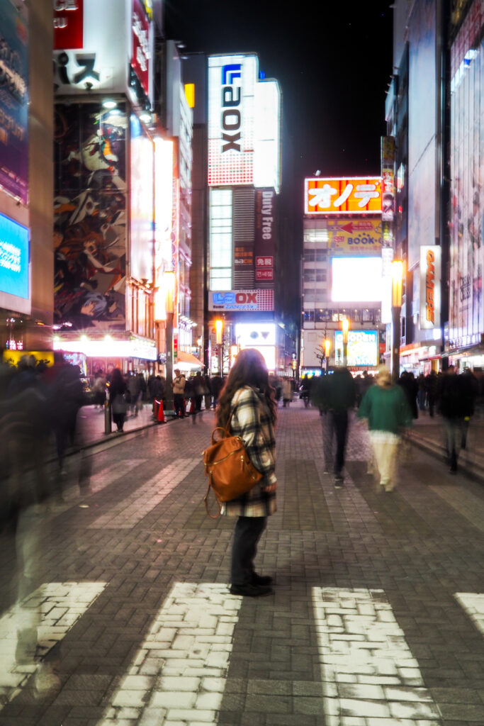 girl standing in the middle of a pedestrian street in Akihabara with people passing by, surrounded by tall lit-up buildings at night