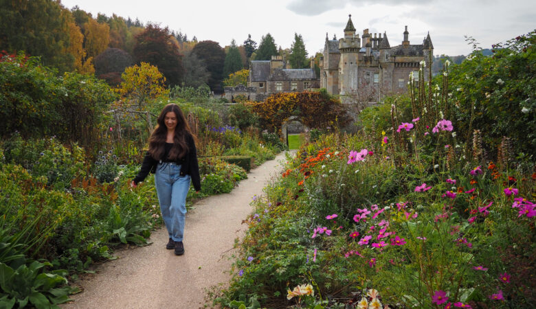 A woman with long dark hair walks along a garden path lined with colorful flowers and greenery, with a large historic castle-like building in the background.