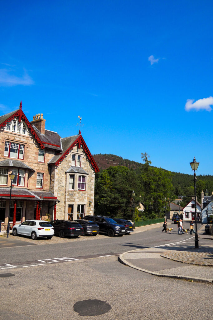 A street scene in a small town with stone buildings, parked cars, a group of people walking, a lamppost, and green hills in the background under a bright blue sky with a few clouds.