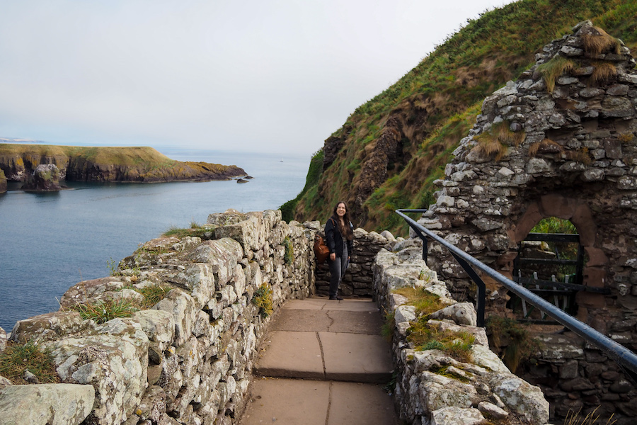 A person stands on a stone pathway among ancient ruins on a hillside, overlooking the sea with green cliffs in the background under a cloudy sky.