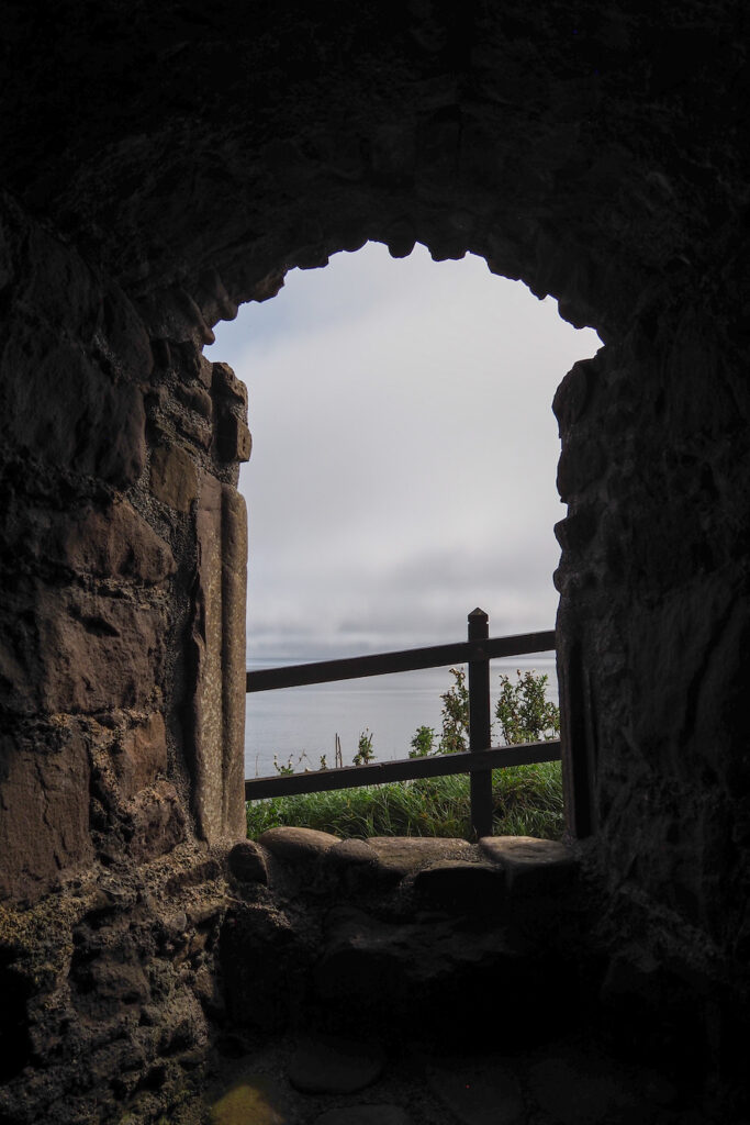 View through a stone archway window, looking out onto a cloudy sky, greenery, and a wooden fence outside. The stone walls are dark, creating a sharp contrast with the light outside.