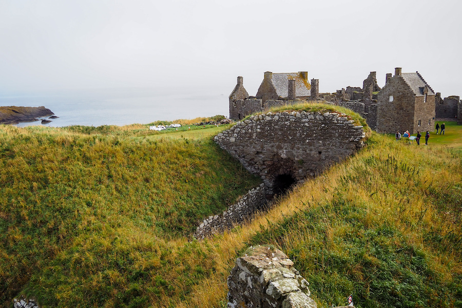 Ruins of a stone castle sit on a grassy hill overlooking the sea under a cloudy sky, with people walking and exploring the area near the old stone structures.