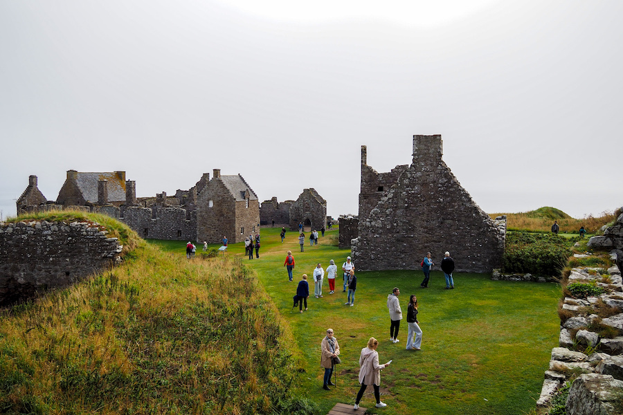 People walk and explore the ruins of old stone buildings surrounded by green grass and low hills, under an overcast sky. The scene appears calm and the ruins suggest a historical site.