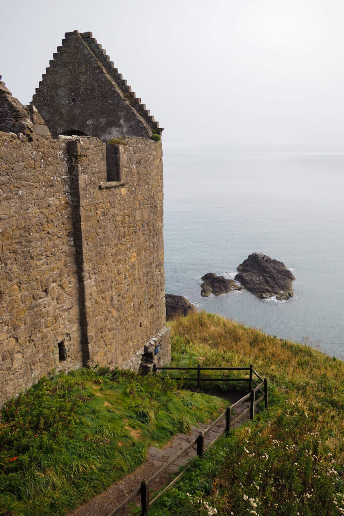 A stone castle wall overlooks a grassy slope and a wooden railing, with rocky outcrops visible in the calm sea below under a hazy sky.