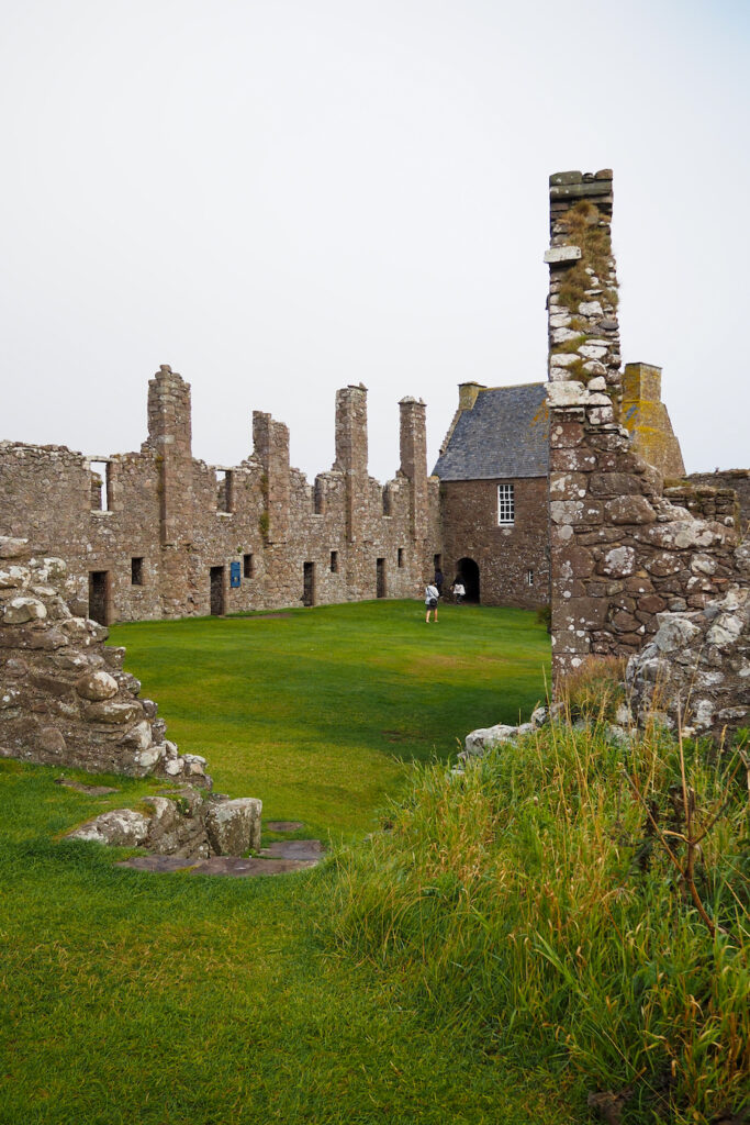 A person stands in the grassy courtyard of an old stone castle ruin, surrounded by tall, weathered walls and towers under a cloudy sky. Some grass and stones are visible in the foreground.