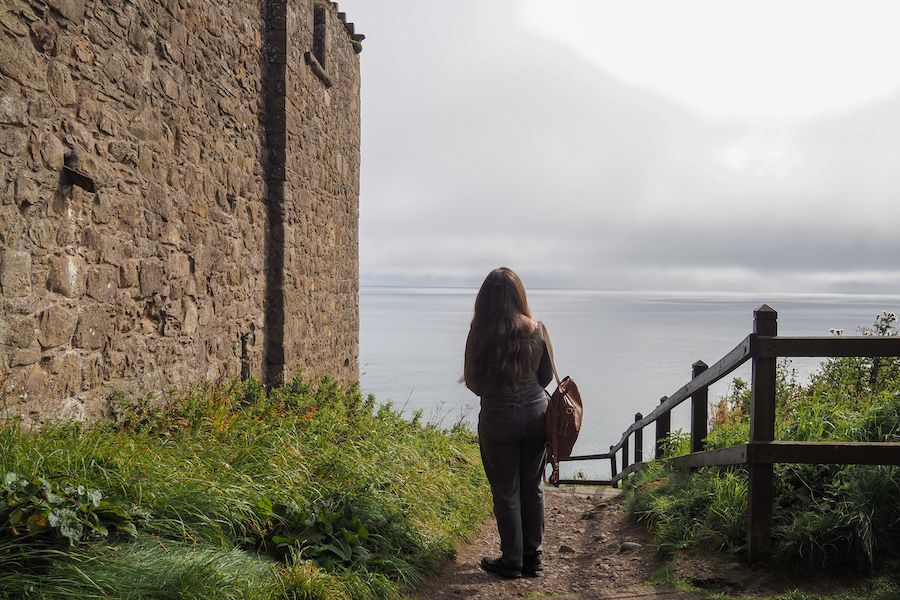 A person with long hair stands near a stone building and wooden fence, looking out over a calm, cloudy sea. Green grass and plants line the path leading to the water.