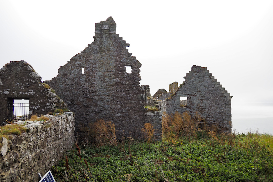 Ruins of an old stone building with gabled walls and empty windows, surrounded by overgrown grass and plants, under a cloudy sky.