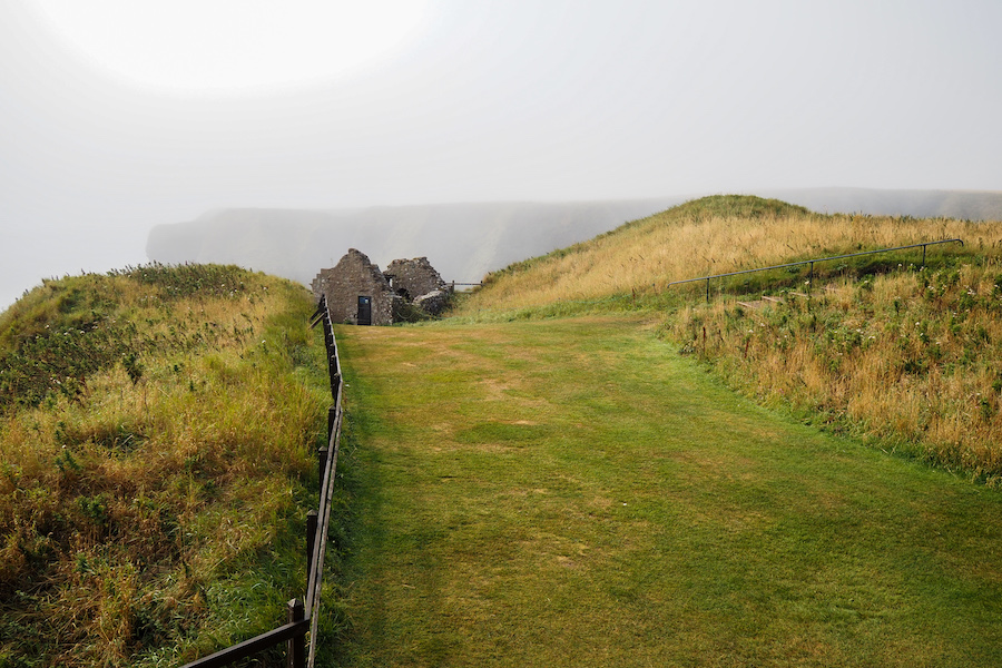 A grassy path leads to stone ruins among green hills, with a wooden fence on both sides. Fog obscures the distant cliffs and sky, creating a serene and slightly mysterious atmosphere.