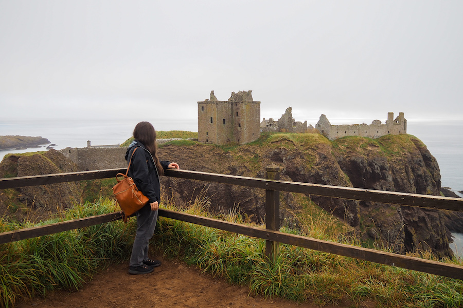 A person with long hair and a brown backpack stands by a wooden fence, looking at the ruins of a cliffside castle overlooking the sea under a cloudy sky.