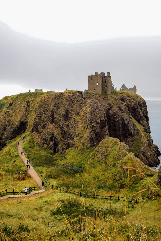Stone ruins of a castle sit atop a steep, grassy cliff overlooking the sea. A narrow path winds up the hill, with people walking toward the historic site under an overcast sky.