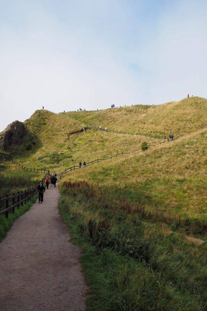 A walking path winds uphill through grassy terrain with people hiking along the trail and scattered on the hill under a pale blue sky.