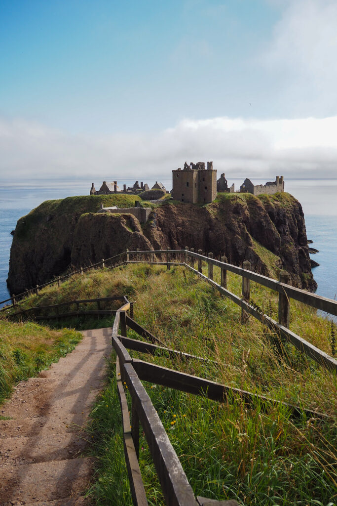 A dirt path with wooden railings leads to the ruins of a stone castle perched on a grassy cliff overlooking the sea under a partly cloudy sky.
