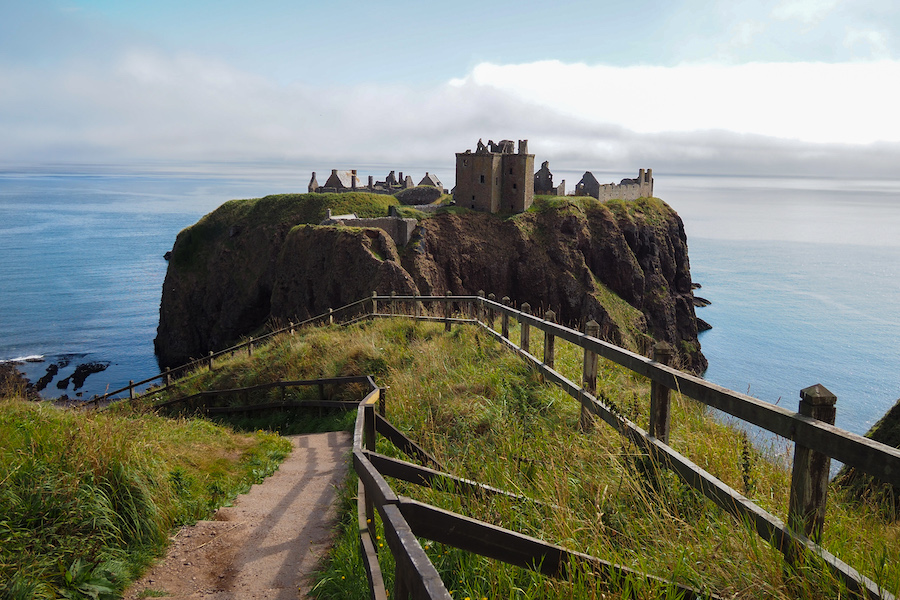 A narrow path with wooden railings leads to the ruins of a medieval castle perched on a grassy cliff overlooking the calm blue sea under a partly cloudy sky.