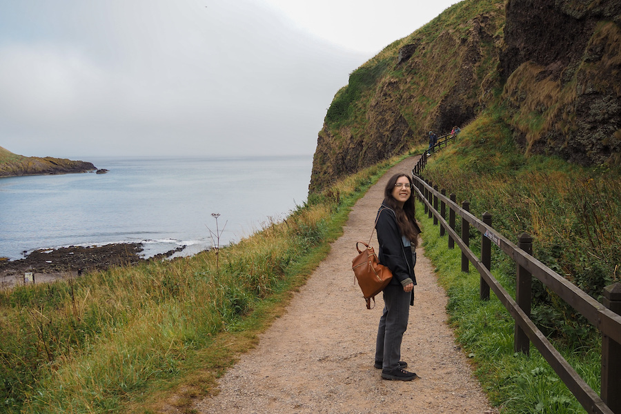 A person with a brown backpack stands on a gravel path beside a grassy hillside, smiling at the camera, with the ocean and rocky shoreline visible in the background.