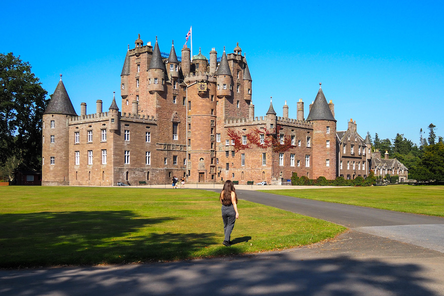 A woman walks across a large, green lawn toward a historic, multi-towered stone castle under a clear blue sky.