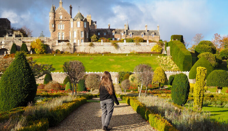 A person with long hair walks on a gravel path through manicured gardens, facing a large stone castle with turrets under a cloudy sky. Neatly trimmed hedges and green lawns surround the scene.