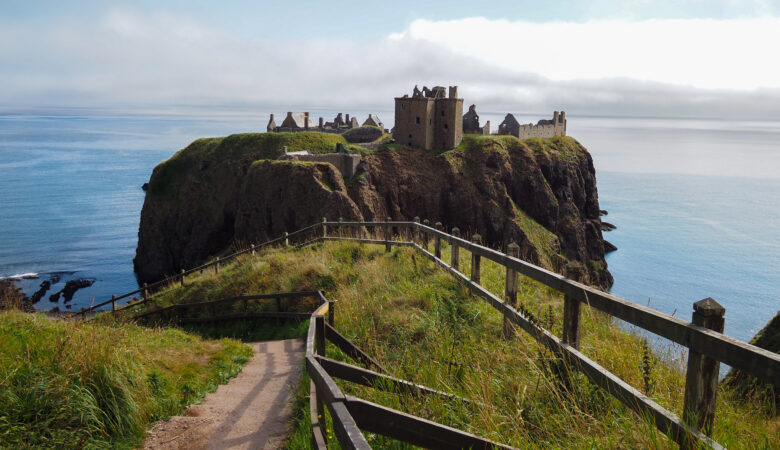 A dirt path with a wooden fence leads to the ruins of Dunnottar Castle, a stone castle, perched on a high cliff overlooking the sea, with grassy slopes and a calm blue ocean in the background.