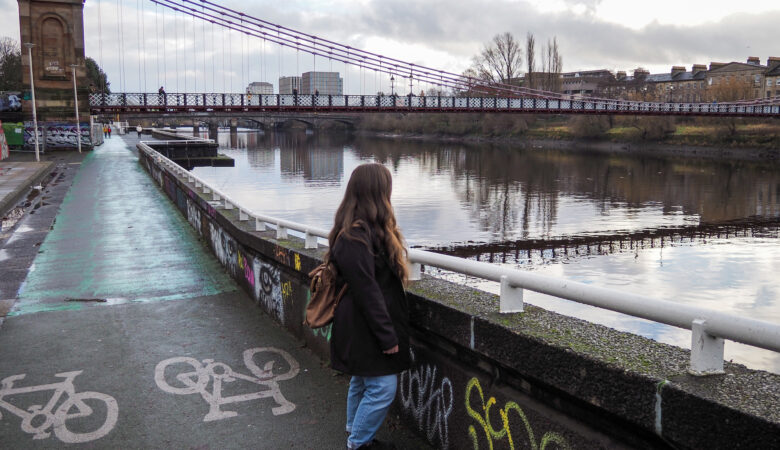 A person with long hair and a backpack stands by a river, looking at a suspension bridge. Bicycle path markings and graffiti are visible on the riverside path. Buildings and trees line the opposite bank.