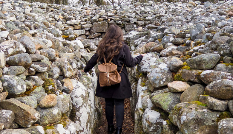 A person with long brown hair and a brown backpack walks through a narrow stone passage surrounded by mossy rocks, with trees visible in the background.