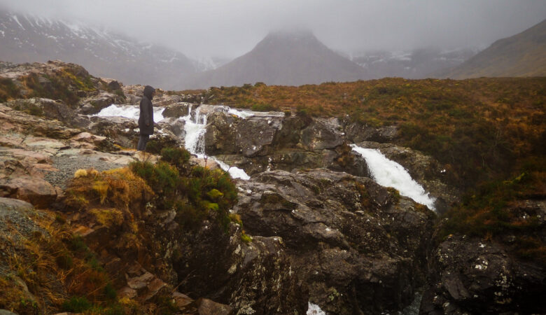 A person in dark clothing stands near rocky waterfalls with misty mountains in the background, under a cloudy sky. The landscape is rugged with patches of grass and flowing water.